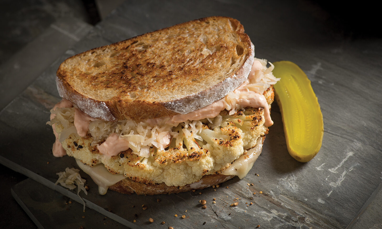 General ACE Bakery® Foodservice Sourdough Bistro with Cauliflower Reuben. Set in a dim black setting with a pickle.